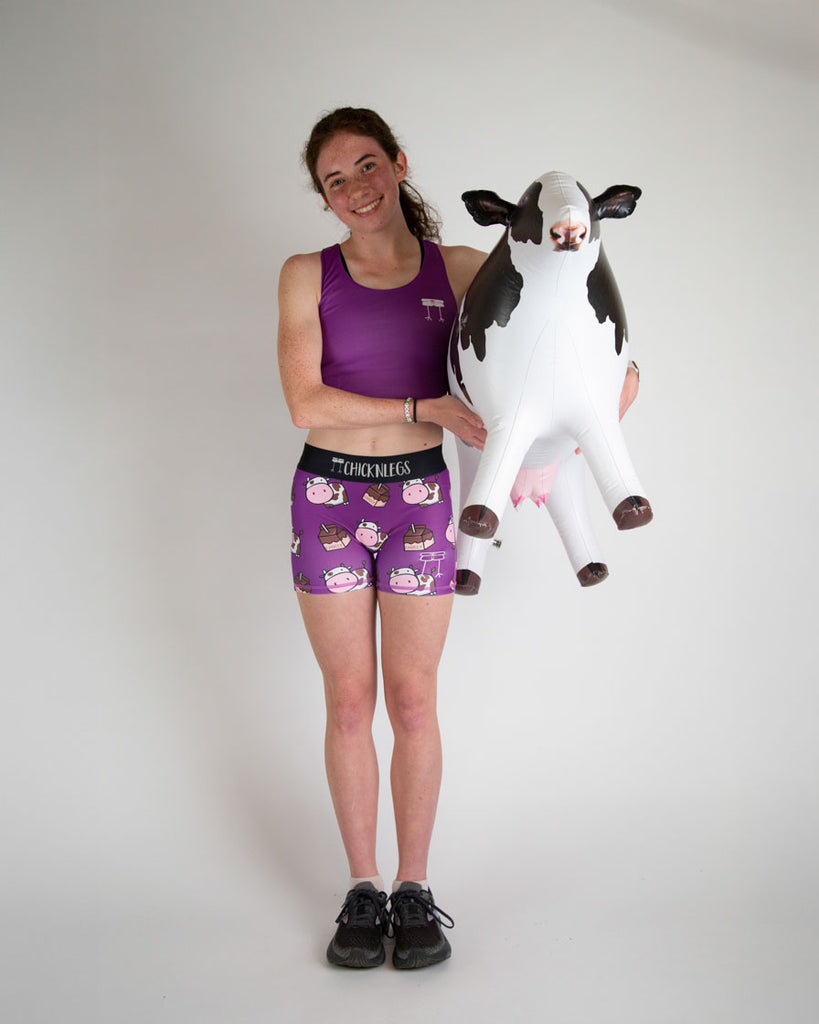 Runner is wearing ChicknLegs in the women's 3 in compression spandex shorts in the choccy cows print. The pattern has chocolate brown cows and chocolate milk on the shorts with a purple background. The model is holding a cow with a huge milk carton next to them.