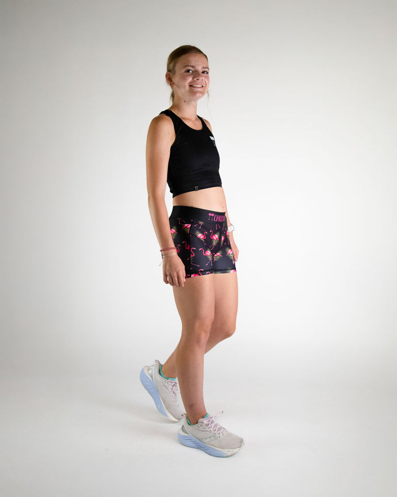 Runner is wearing ChicknLegs in the women's 3 in compression spandex shorts in the black flamingo design. The print has bright pink flamingos on it with green leaves on a black background. Full body image is shown of the model to show the matching fit with wearing the flamingos with the black crop top.