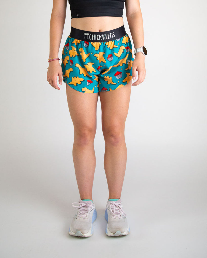 Runner is wearing ChicknLegs in the women's 4 in split running shorts in the dino nuggets design. The print has dinosaur chicken nuggets on it with ketchup on a blue background. 