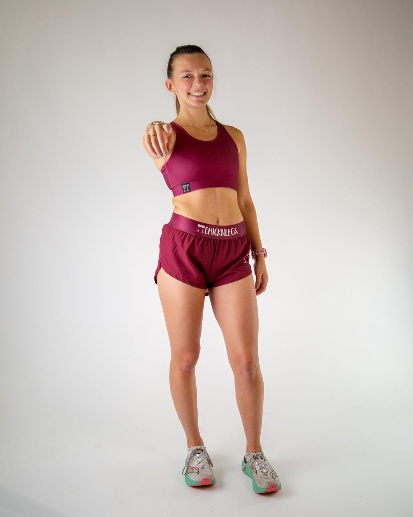 Full body view of model wearing ChicknLegs women’s og sports bra in solid maroon, styled with maroon running shorts and white running shoes.