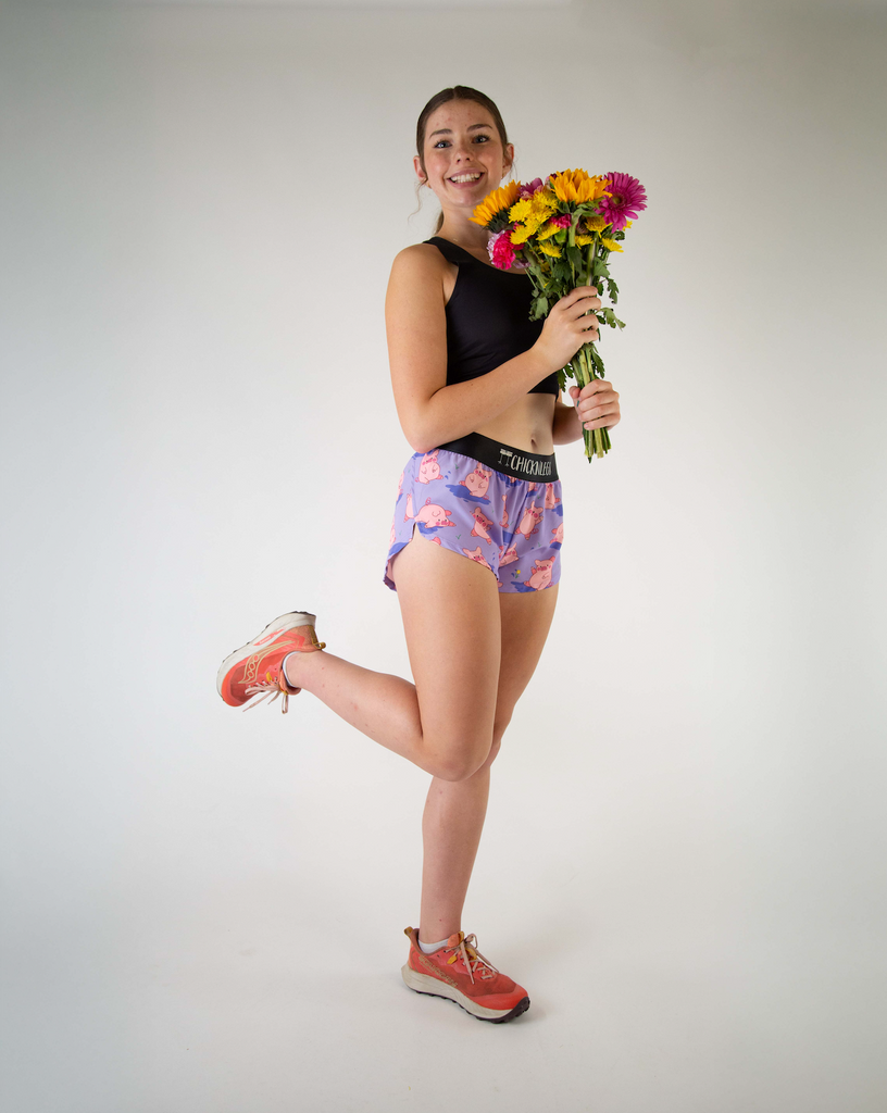 Full body view of ChicknLegs women’s 1.5-inch split running shorts with drawstring, breathable liner, lilac purple background and pink piggies design. Runner is holding a bouquet of flowers.