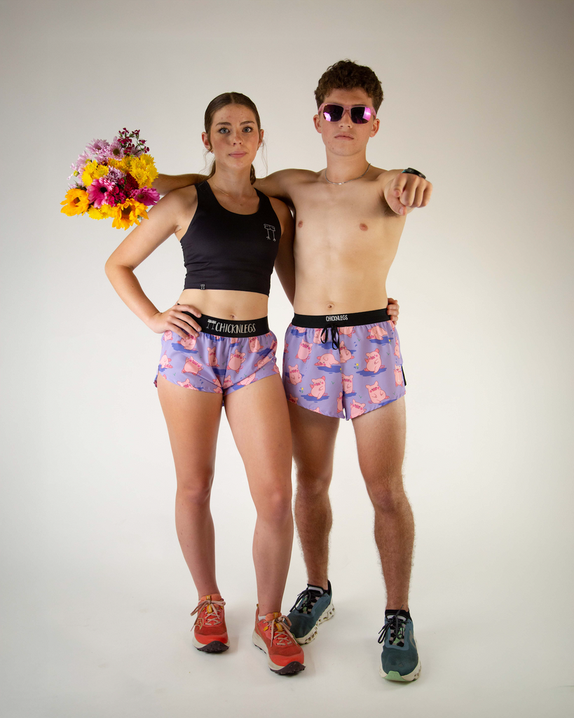 Duo photo of ChicknLegs men’s 4-inch split running shorts, breathable liner, with women's 1.5 inch split running shorts with a lilac purple background and pink piggies on the design. The runners are wearing sunglasses and holding flowers.