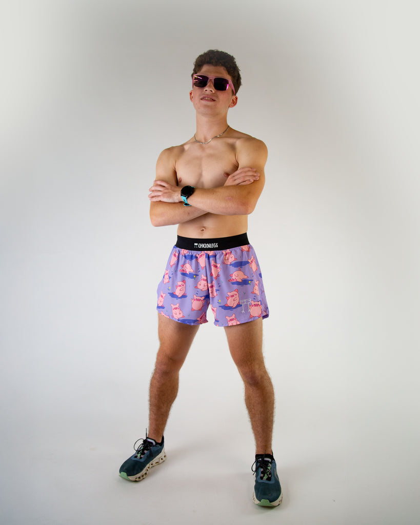 Full body view of ChicknLegs men’s 4-inch split running shorts, breathable liner, lilac purple background and pink piggies on the design. Paired with pink sunglasses.