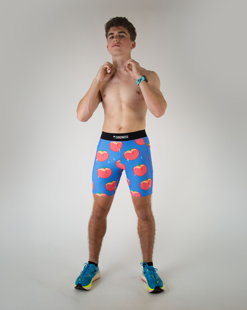 Full body view of ChicknLegs men’s 8-inch half tights, breathable liner, black waistband and blue background and juicy peaches design. Side pockets are visible in the photo, paired with blue shoes.