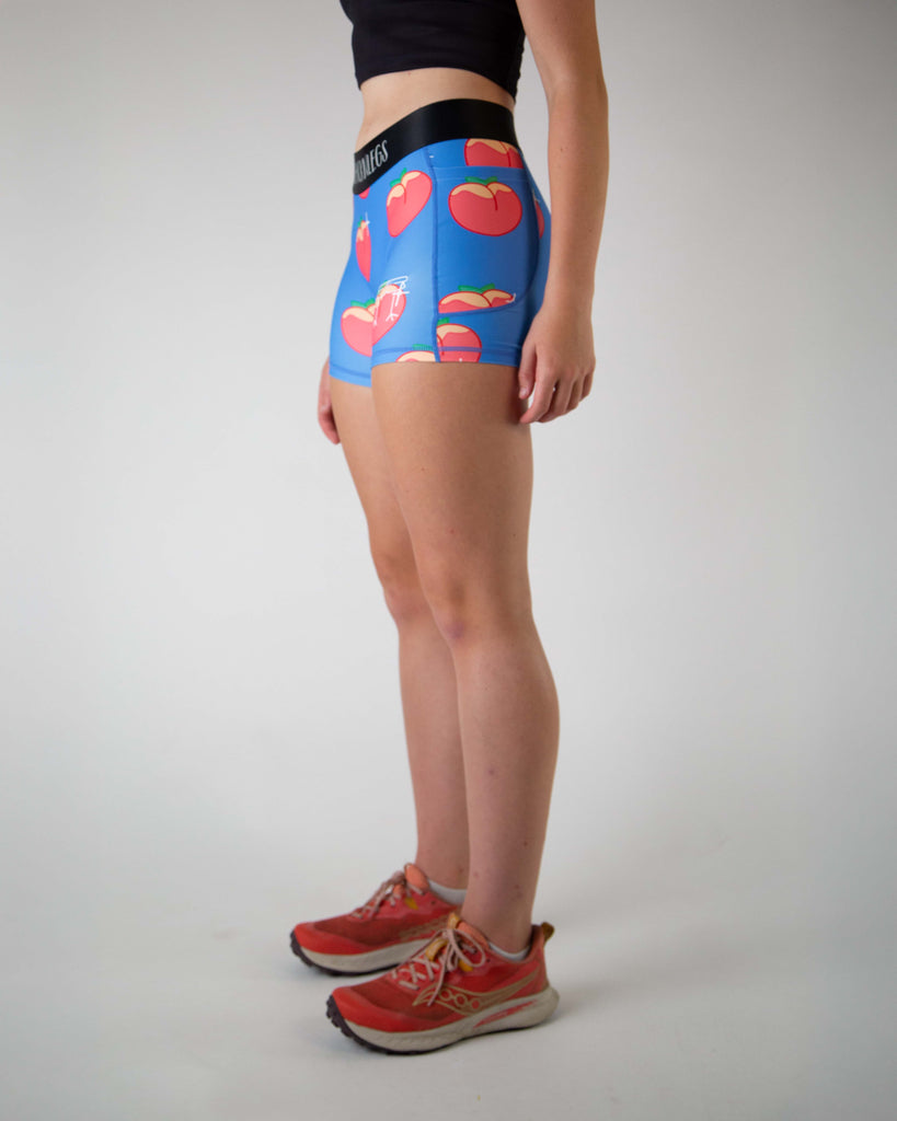 Left side view of ChicknLegs women’s 3-inch compression spandex shorts, black non chaffing waistband, blue background and juicy peaches on the design, with side pockets visible.