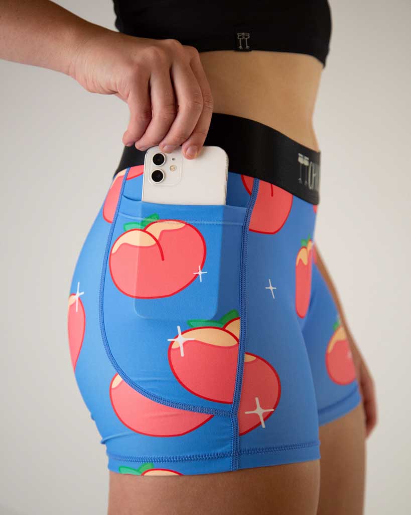 A close up shot view of ChicknLegs women’s 3-inch compression spandex shorts, black non chaffing waistband, blue background and juicy peaches on the design. The runner is holding a phone in the pocket.