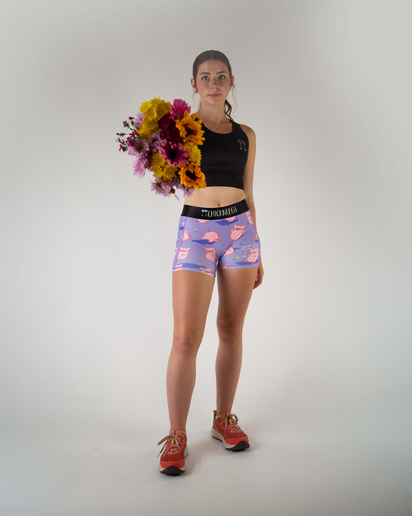 Full body view of ChicknLegs women’s 3-inch compression spandex with side pockets, lilac purple background and pink piggies design, runner is holding flowers to pair with pigs.