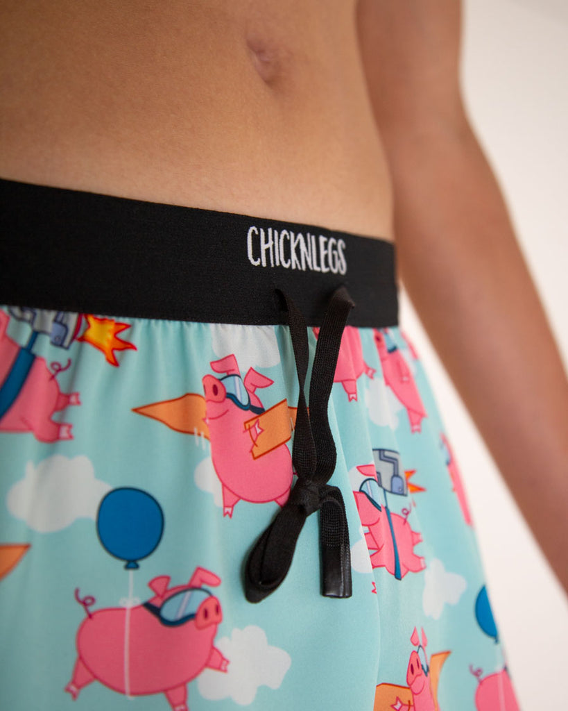 A close up image is shown of ChicknLegs men's 2 in drawstrings split running shorts in the flying pig design. The print has pink piggies all over it in different ways that they are flying.