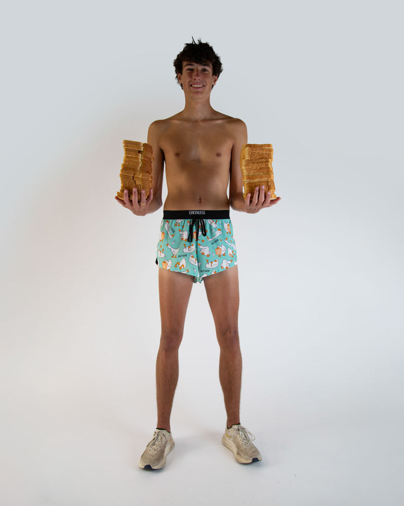 Runner is wearing ChicknLegs in the men's 2 in drawstring split running shorts in the Silly Goose design. The model is holding loafs of bread in various piles, rye, wheat, whole bread, white.