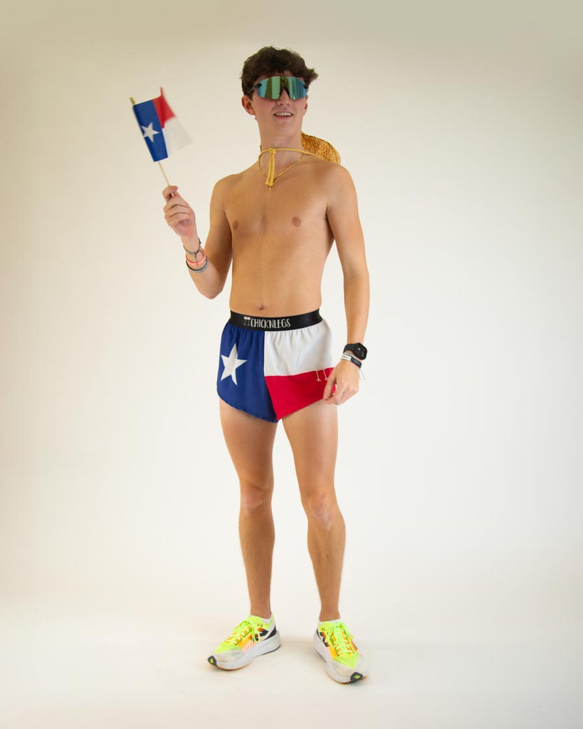 Model is wearing ChicknLegs in the men's 2 in split running shorts n the texas flag design, wearing a cowboy hat and waving a flag.