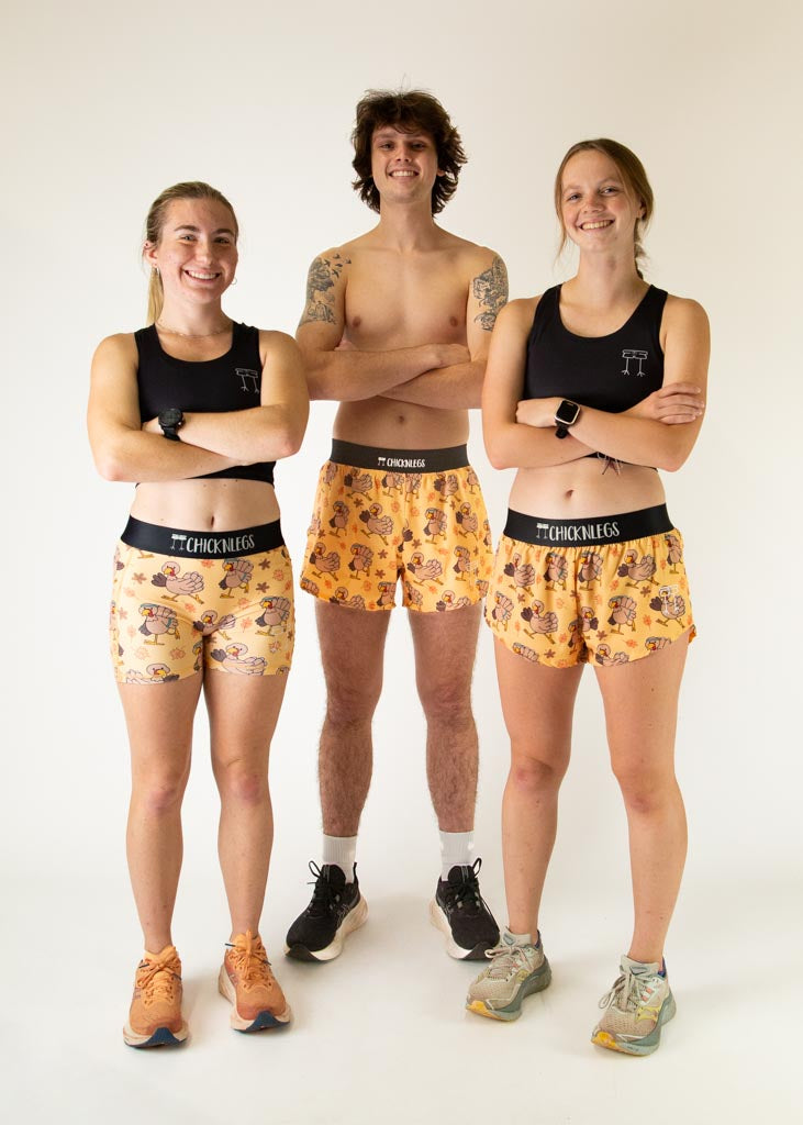 The model is a runner who is wearing Chicknlegs men's 4 inch split running shorts in the pardoned turkey design, facing front while posing with two other models who are runners wearing the women's 3 inch compression shorts and women's 1.5 inch split running shorts in the pardoned turkey design.