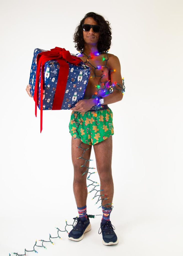 The model is a runner who is wearing Chicknlegs men's 2 inch split running shorts in the Gingerbread Men's Gingy Revenge design, facing front wrapped up in christmas lights and holding a giant present.