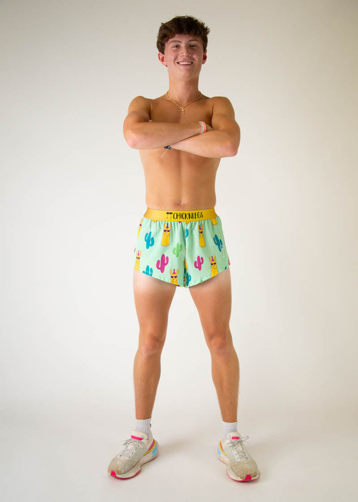 The model is a runner wearing Chicknlegs in men's 2 inch split running shorts in the green llama design, facing front while posing and crossing their arms.