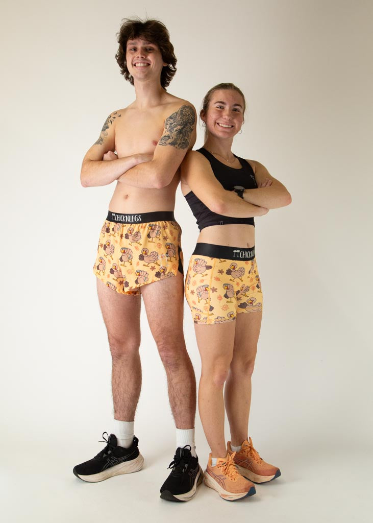 The model is a runner who is wearing Chicknlegs in men's 2 inch split running shorts in the pardoned turkey design, facing front and posing with another model who is a runner and wearing the women's 3 inch compression pardoned turkey shorts.