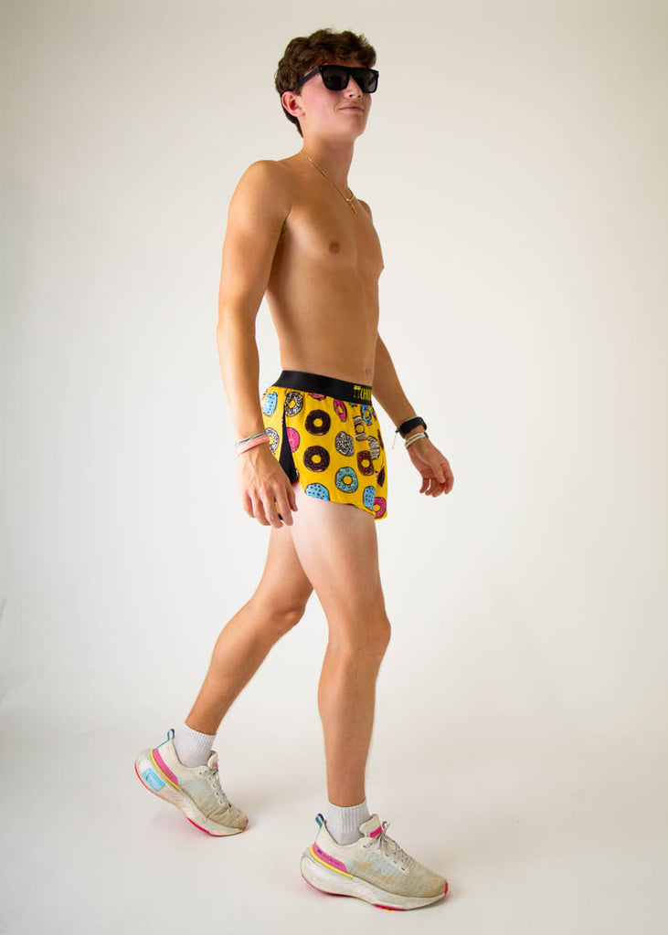 The model is a runner that is wearing Chicknlegs men's 2 inch split running shorts in the salty donuts design, posing in a walking stance facing right.