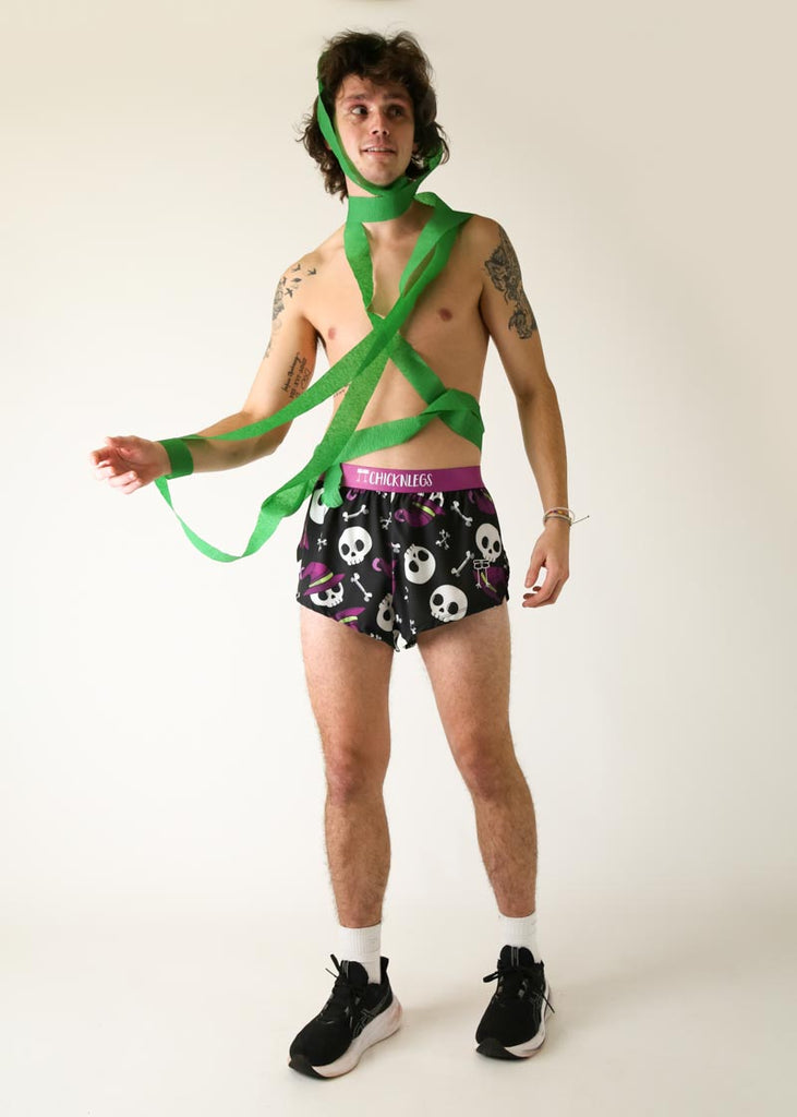 Model is a runner wearing Chicknlegs men's 2 inch split running shorts in the Skulls design, posing while being scared and holding green streamer.