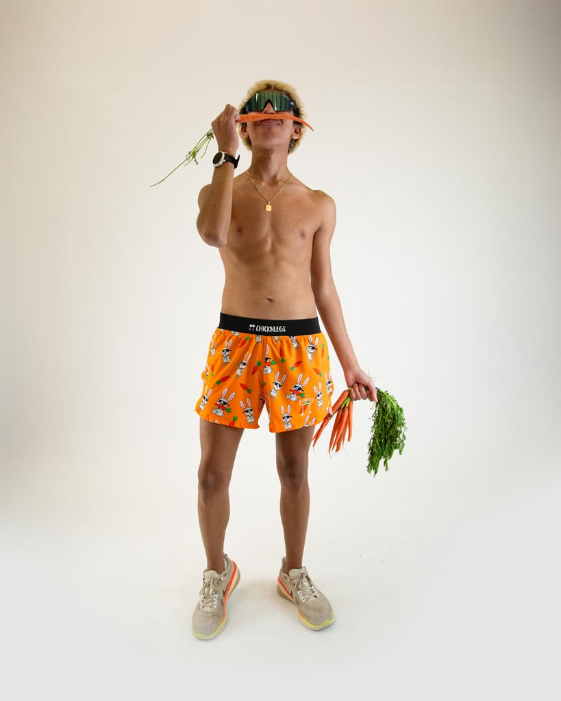 Runner is wearing ChicknLegs in the men's 4 in split running shorts in the bad bunny design, facing front. The print has white rabbits on it with sunglasses and carrots on an orange background. The model is posing with real vegetables and carrots.