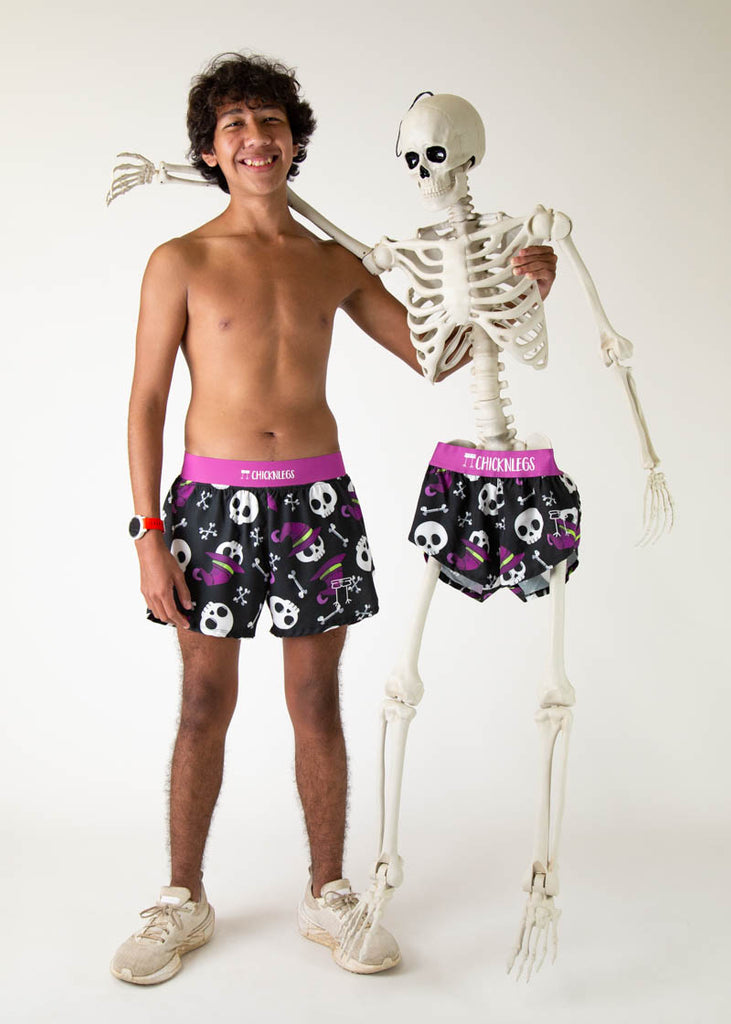 The model is a runner who is wearing Chicknlegs running shorts in the men's 4 inch split runners shorts in the skulls design, facing front and posing with a skeleton.