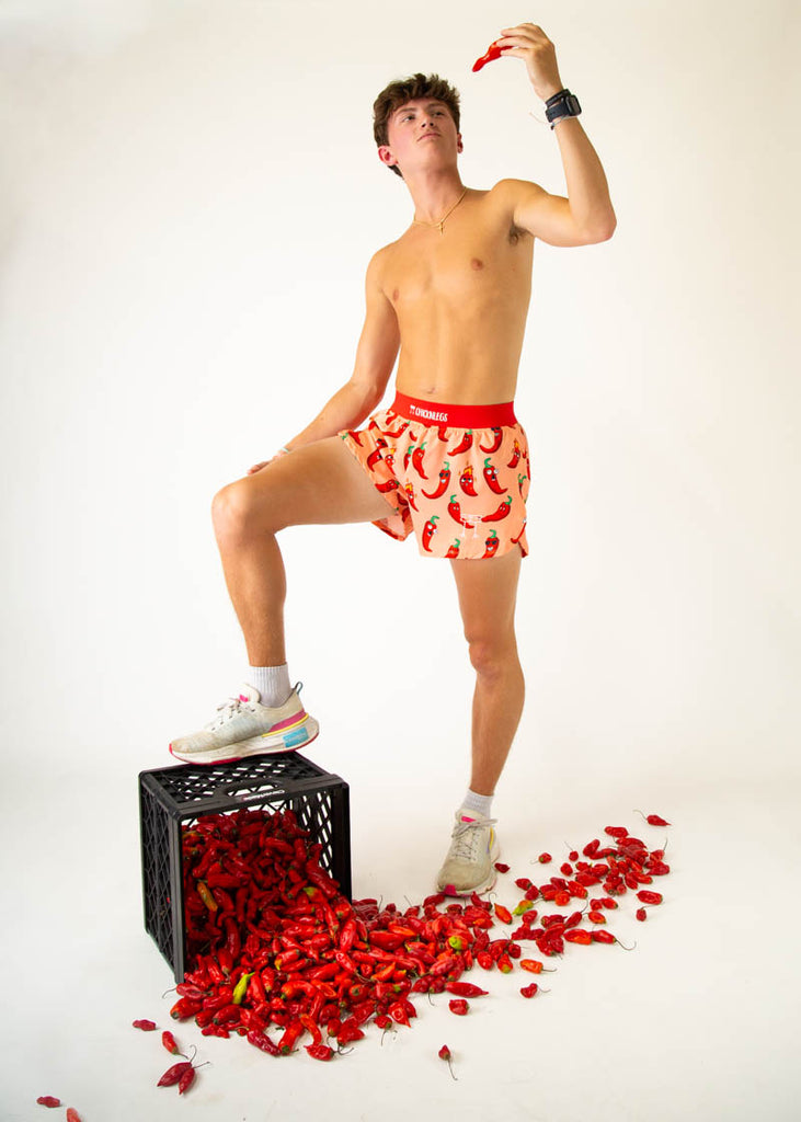 Model is a runner wearing Chicknlegs 4 inch split running shorts in the Chili Pepper design, facing front while posing with chili peppers.