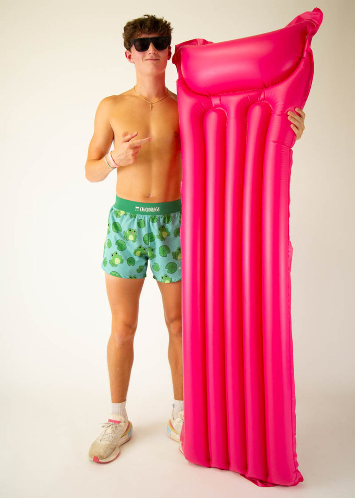 Model is a runner wearing Chicknleg's men's 4 inch split running shorts in the fartlek frog design, facing front while posing with a  floatie to show the shorts.
