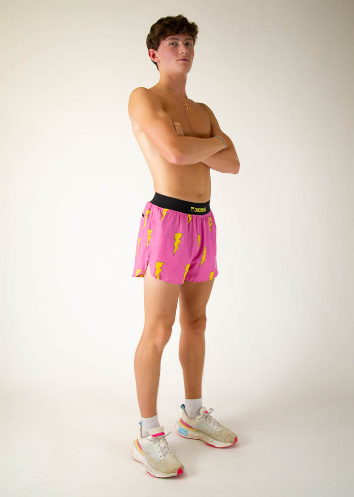 The model is a runner wearing Chicknlegs men's 4 inch split running shorts in the hot pink bolts design, facing right and crossing their arms looking at the camera.