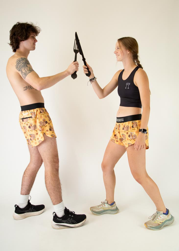 The model is a runner who is wearing Chicknlegs women's 1.5" split running shorts in the pardoned turkey design, posing with another model who is a runner wearing the men's 4 inch split running shorts in the pardoned turkey design.