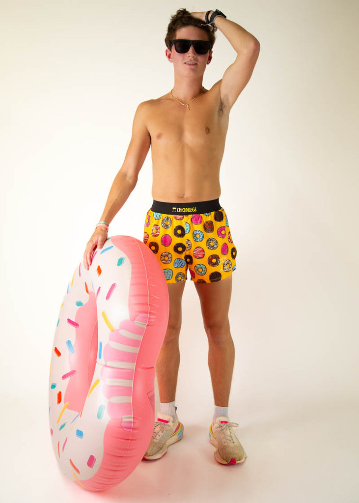 The model is a runner wearing Chicknlegs in the men's 4 inch split running shorts in the salty donuts design, facing front while holding a giant donut.