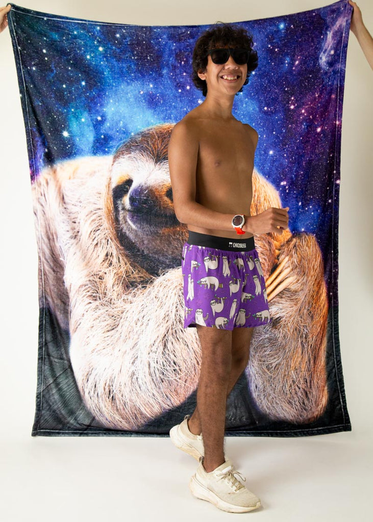 The model is a runner who is wearing Chicknlegs men's 4 inch split running shorts in the sloth design, facing front in a running pose while a giant picture of a sloth is hung and held up in the background.