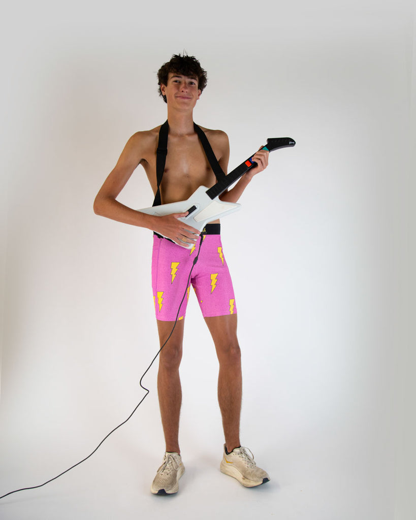 Runner is wearing ChicknLegs in the men's 8 in half tights in the neon hot pink lightning bolts. The print is on bright vibrant pink with yellow bolts. Runner is holding an electric guitar. 