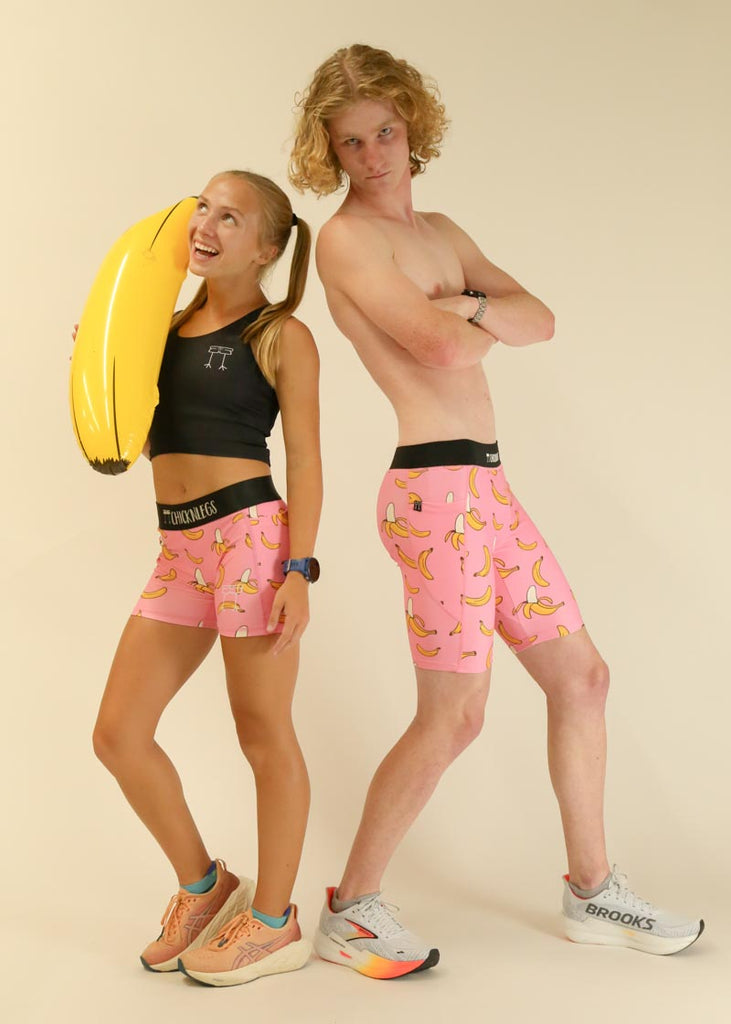 Model wearing Chicknlegs men's 8 inch half tights in the pink bananas design along with another model wearing women's 3 inch compression shorts in the pink bananas design, both posing together with a banana.