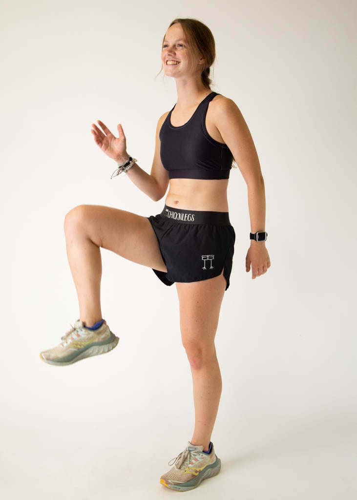 The model is a runner wearing Chicknlegs women's OG sports bra in the black design, posing in a high knee stance.