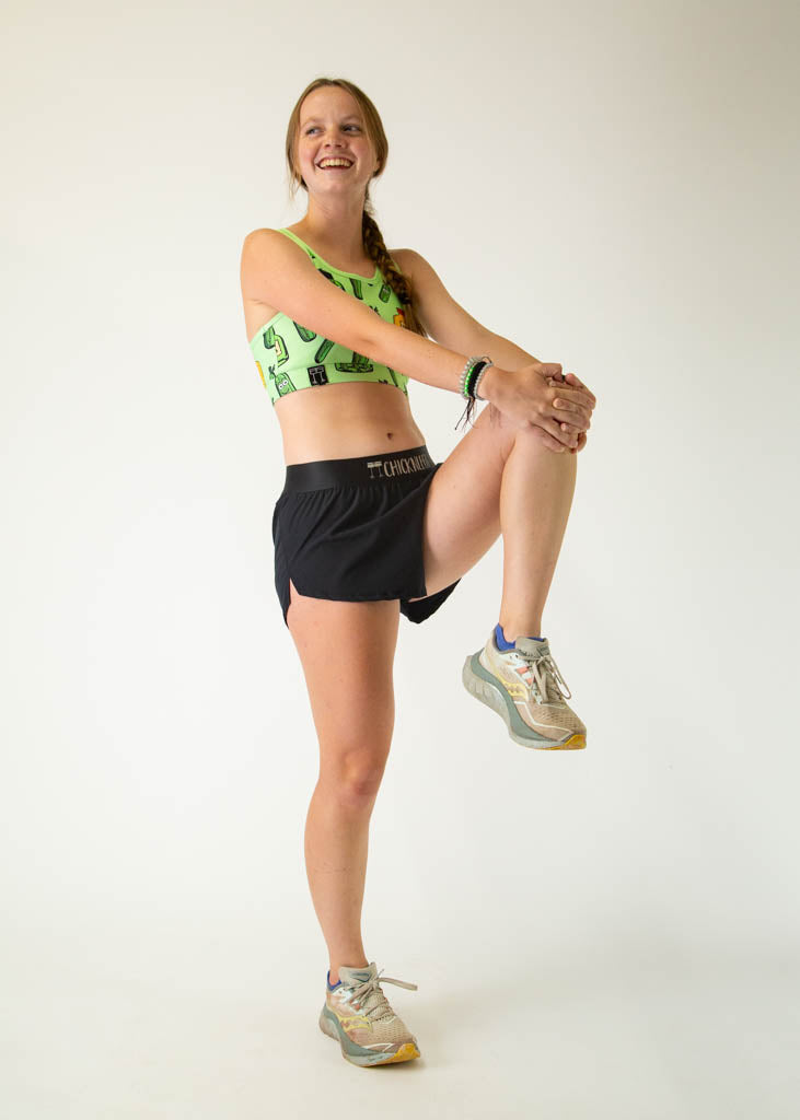 The model is a runner who is wearing Chicknlegs women's OG sports bra in the pickles design, facing front while stretching their quad, a full body pose is shown.
