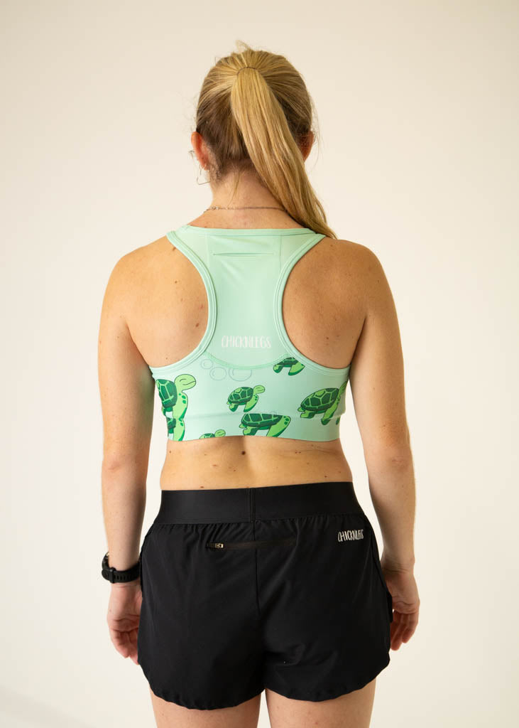 The model is a runner who is wearing Chicknlegs OG sports bra in the turtles design, facing away from the camera to show the back of the sports bra, a pocket is included.