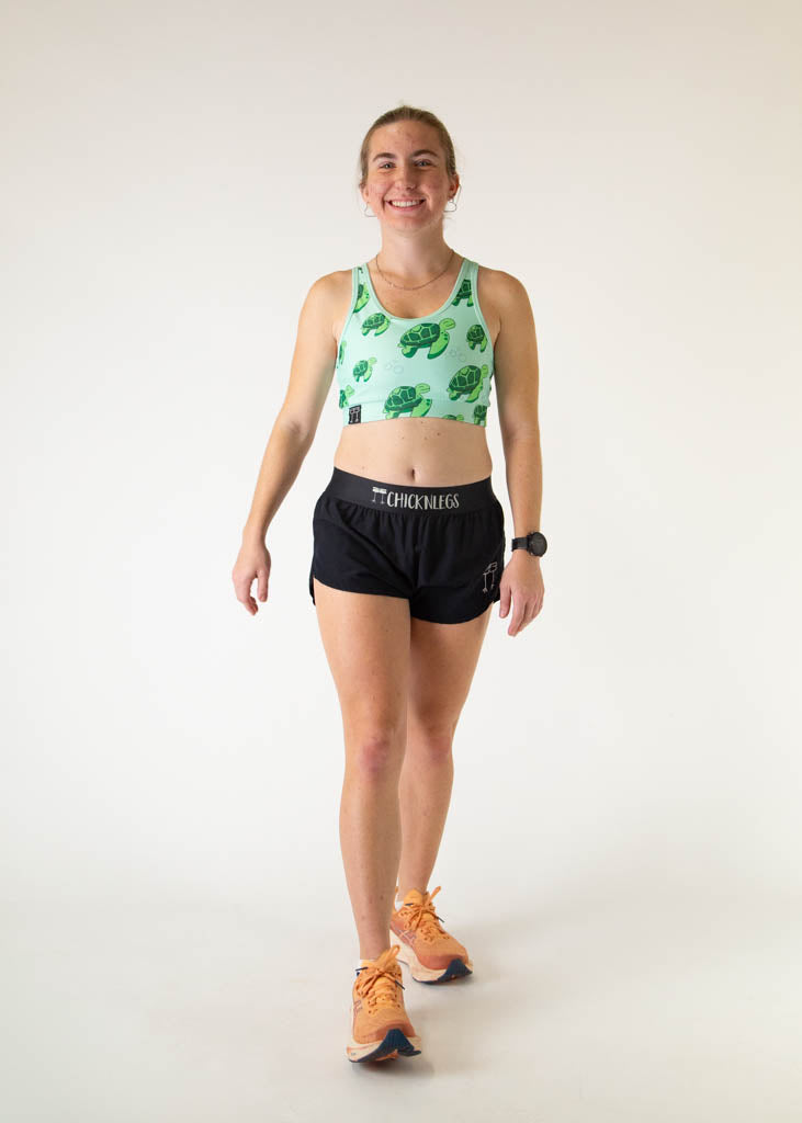 The model is a runner who is wearing Chicknlegs OG sports bra in the turtles design, facing front posing in a walking stance. 