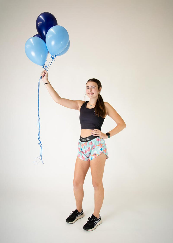 Runner is wearing ChicknLegs in the women's 1.5 in split running shorts in the flying pigs design, facing front. The design has pigs with cardboard, balloons, wings and jetpacks with clouds the animal print is fun and cute, holding huge dark blue balloons and light blue balloons.