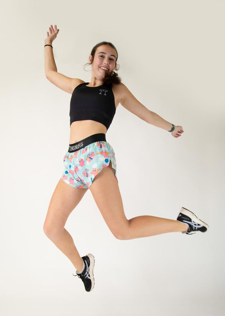 Runner is wearing ChicknLegs in the women's 1.5 in split running shorts in the flying pigs design, facing front. The design has pigs with cardboard, balloons, wings and jetpacks with clouds. Model is jumping high to fly with pigs.