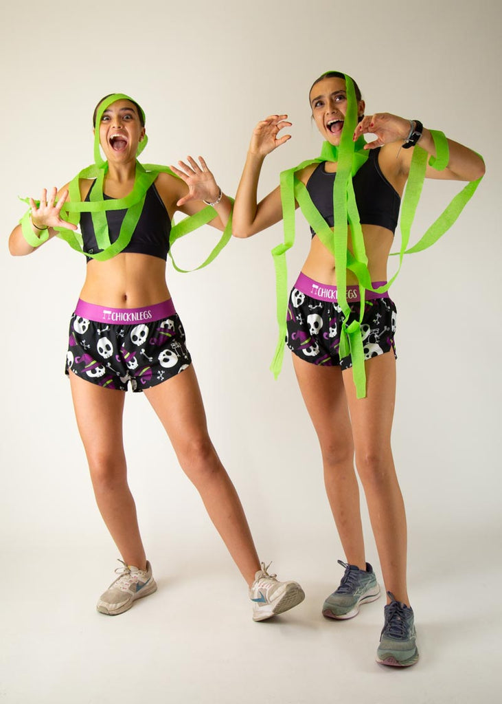 Two models who are runners are posing in the Chicknleg's women's 1.5" split running shorts in the Skulls design, posing while looking scared.