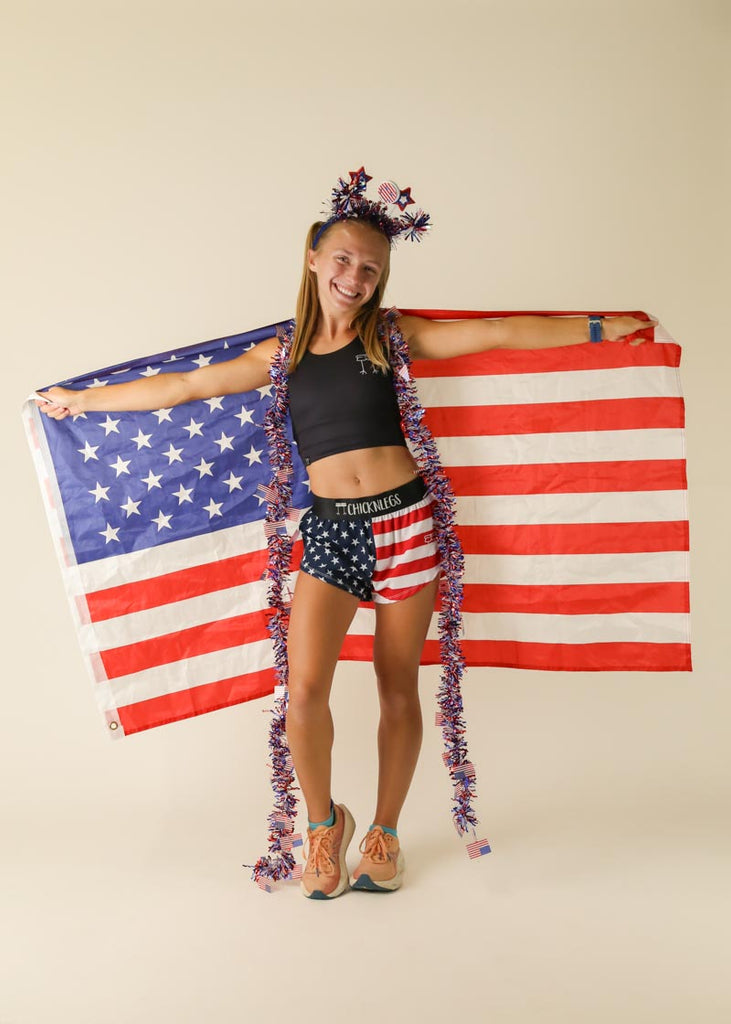 Model is wearing Chicknlegs women's 1.5 inch split running shorts in the USA design posing with the american flag and usa tinsel. 
