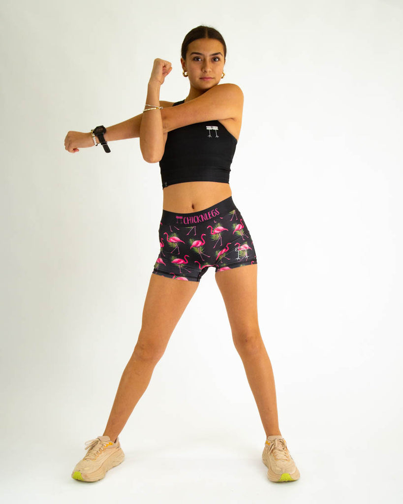 Runner is wearing ChciknLegs in the women's 3 in compression spandex shorts in the black flamingo design. Model is posing while stretching and wearing a matching top with the black pwr crop top.