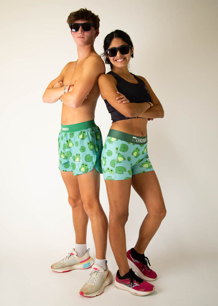 Model is a runner wearing Chicknlegs 3 inch compression shorts in the fartlek froggies frog design, while also posing with a male model who is a runner, posing and wearing the men's 4 inch split running shorts in the fartlek froggies frog design, facing front while wearing sunglasses and crossing their arms.
