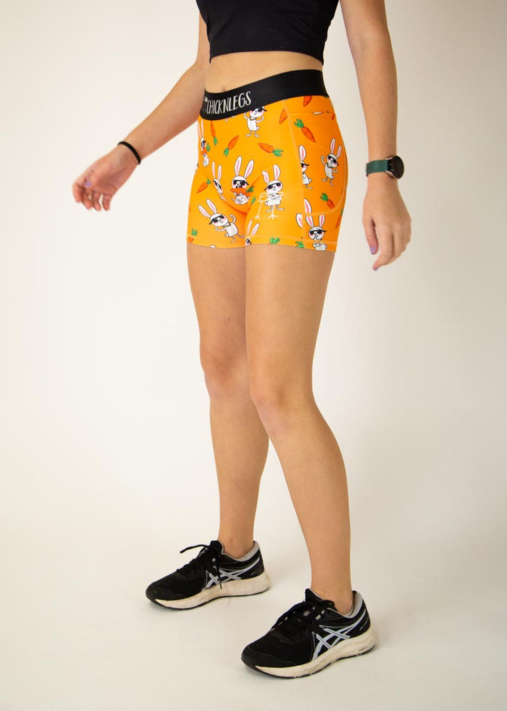 Runner is wearing ChicknLegs in the women's compression spandex in the bad bunny design, facing left. The print has white cute hare on it with sunglasses and carrots flexing their muscles on an orange background.