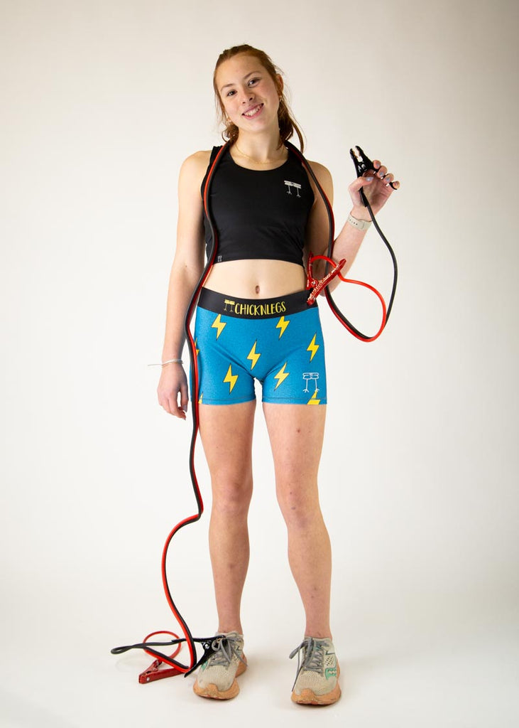 The model is a runner who is wearing Chicknlegs in the women's 3" compression shorts in the blue bolts design, facing front while posing with jumper cables.