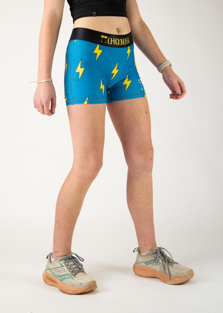 The model is a runner who is wearing Chicknlegs in the women's 3" compression shorts in the blue bolts design, facing right.