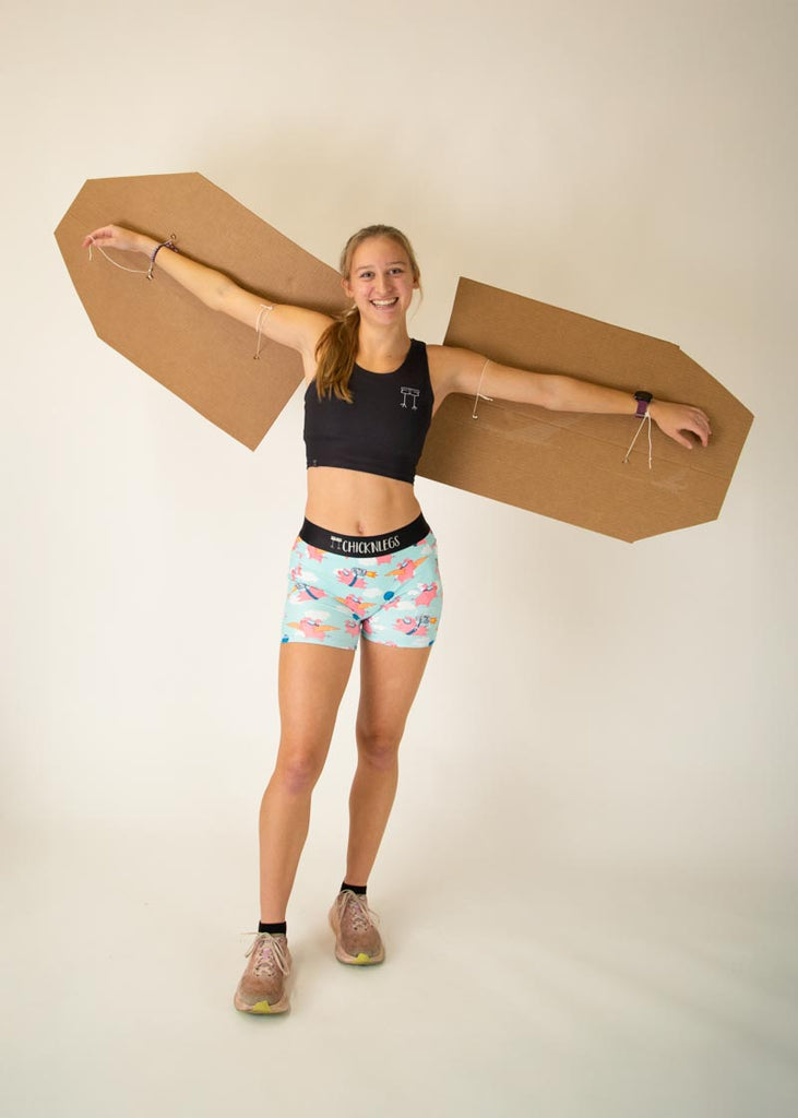 Runner is wearing ChicknLegs in the women's 3 in compression spandex shorts with side panel pockets that fit high rise on the waist. The print has flying pigs on it with clouds.The model is wearing giant cardboard wings.