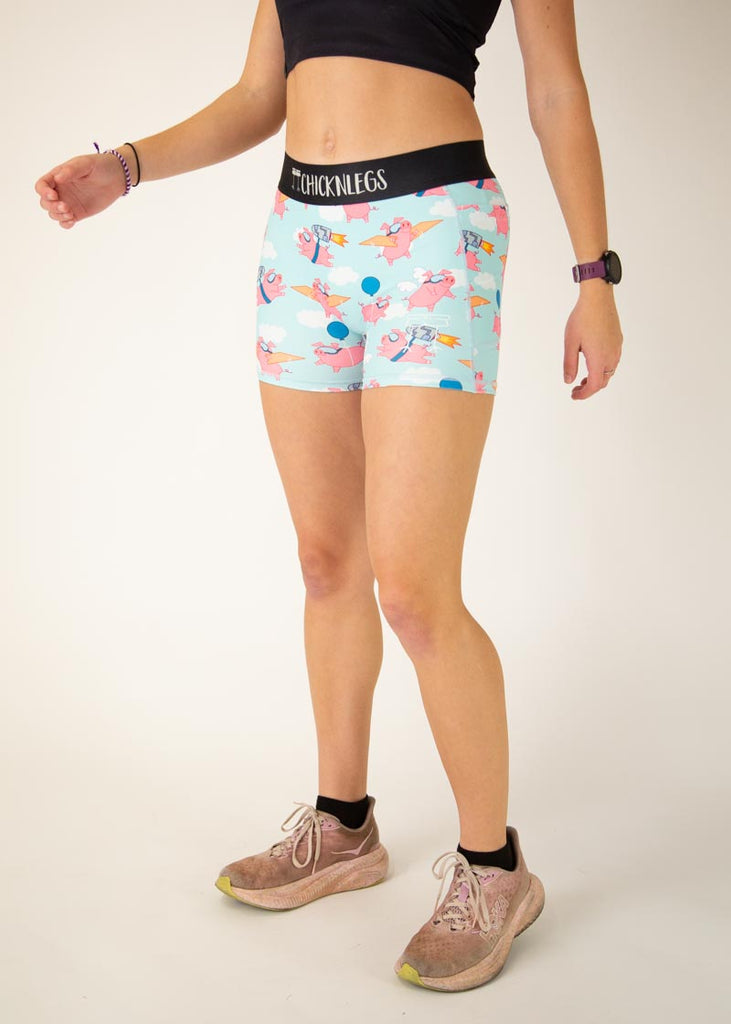 Runner is wearing ChicknLegs in the women's 3 in compression spandex shorts with side panel pockets that fit high rise on the waist. The print has flying pigs on it with clouds and blue balloons.