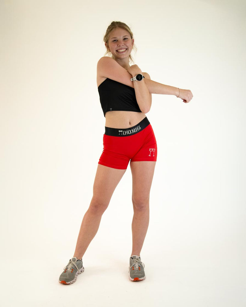 Runner is wearing ChicknLegs in the women's 3 in compression spandex shorts in the solid red design. Model is posing while stretching her arm.