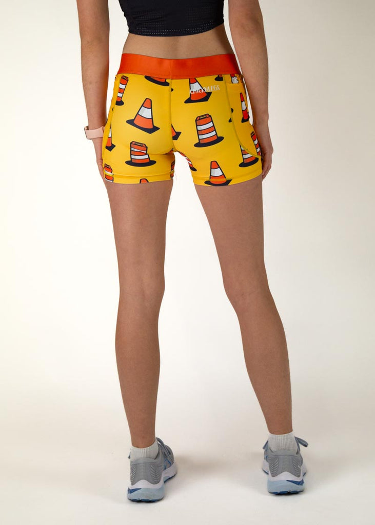 The model is a runner who is wearing Chicknlegs in women's 3" compression shorts in the traffic cones design, facing away from the camera to show the back of the compression.