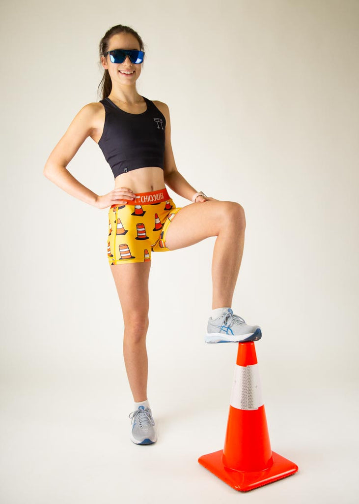 The model is a runner who is wearing Chicknlegs in women's 3" compression shorts in the traffic cones design, facing front while posing for a full body photo with sunglasses and a traffic cone.