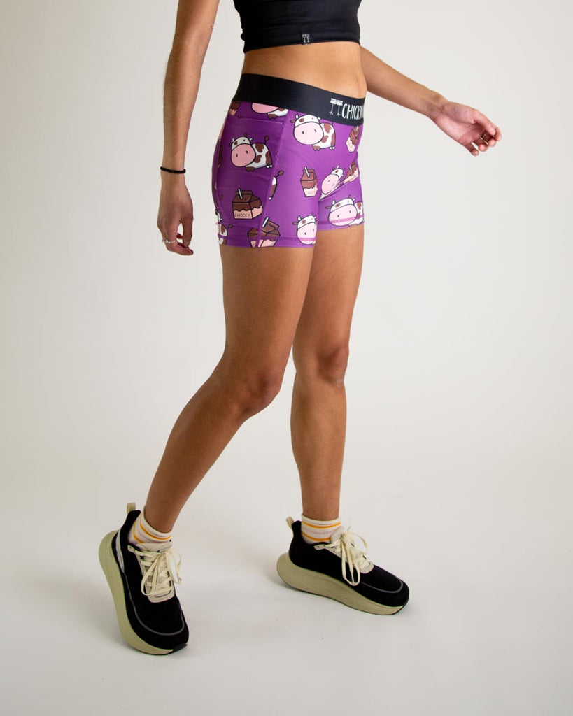 Runner is wearing ChicknLegs in the women's 3 in compression spandex shorts with pocket in the choccy cows design. The print has brown and white chocolate cows with milk cartons on it. The model is facing right.