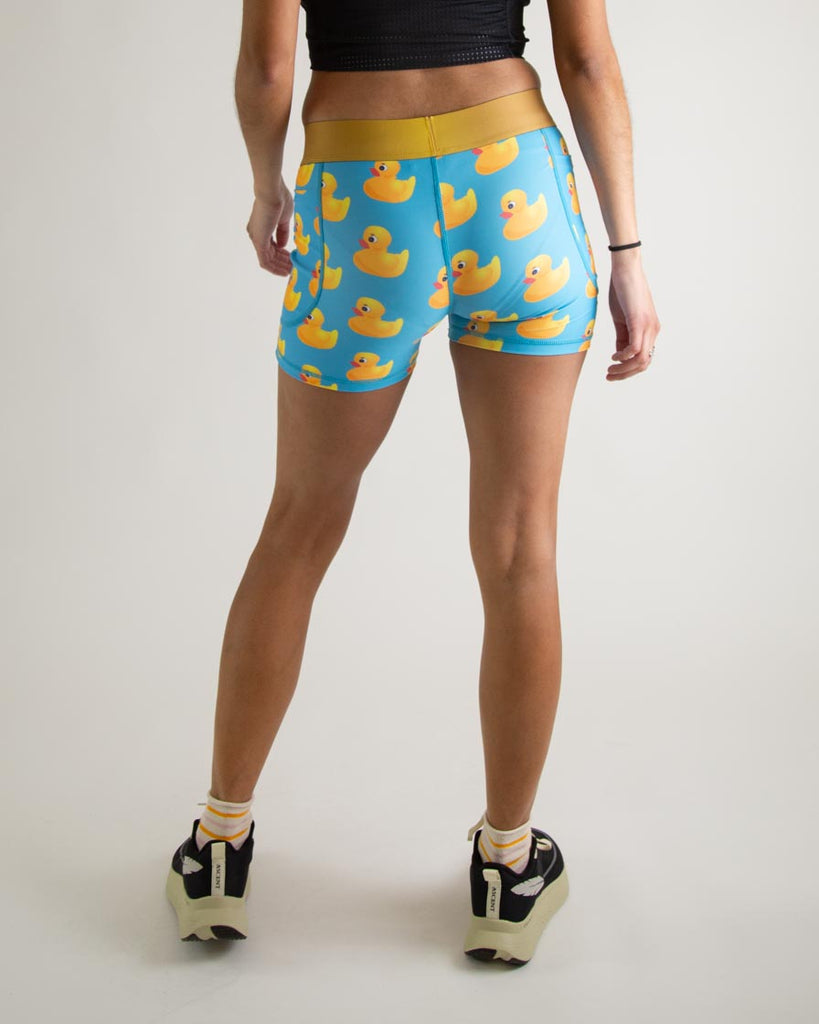 Runner is wearing ChicknLegs in the women's 3 in compression spandex shorts with pockets in the rubber ducky design. The print has yellow rubber ducks on it with a blue background. The model is facing away from the camera to show the back.